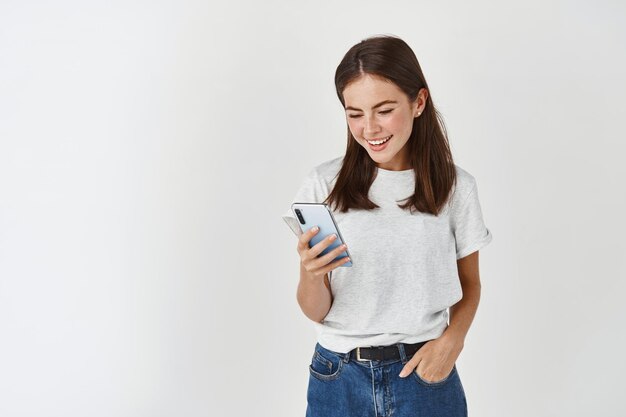 Jonge mooie dame leest bericht op smartphone, kijkt naar mobiel scherm en glimlacht, staande in vrijetijdskleding over witte muur
