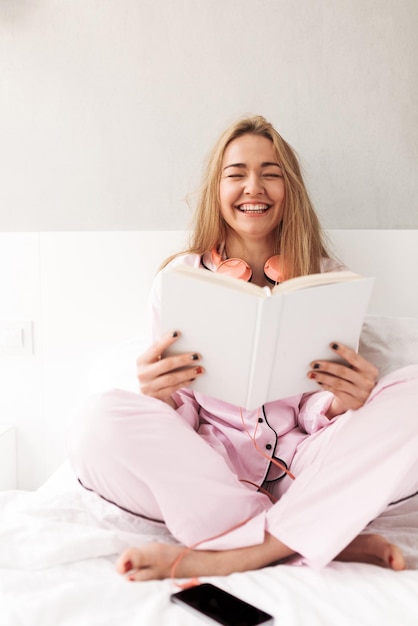 Jonge mooie dame zittend op bed met boek in handen en thuis lachen