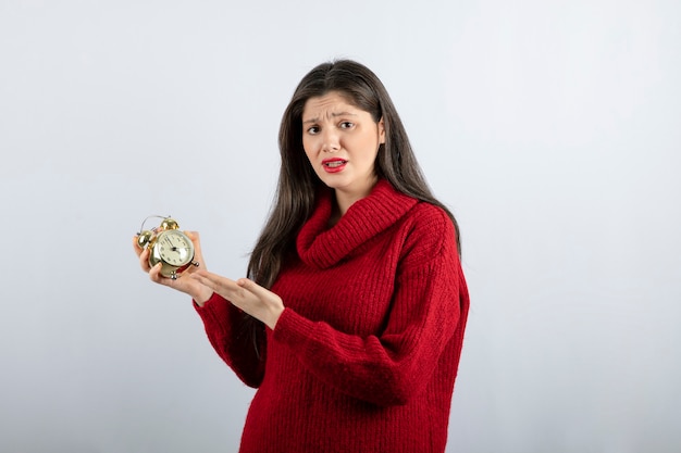 Jonge mooie donkerbruine vrouw die een wekker houdt die zich over witte achtergrond bevindt