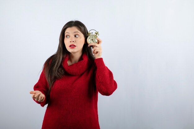 Jonge mooie donkerbruine vrouw die een wekker houdt die zich over witte achtergrond bevindt