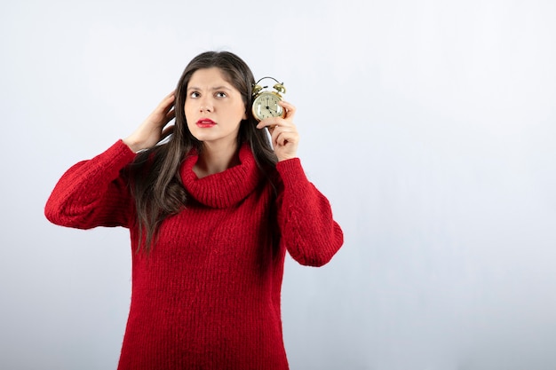 Jonge mooie donkerbruine vrouw die een wekker houdt die zich over witte achtergrond bevindt