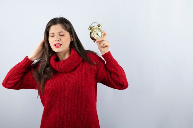 Jonge mooie donkerbruine vrouw die een wekker houdt die zich over witte achtergrond bevindt