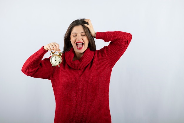 Jonge mooie donkerbruine vrouw die een wekker houdt die zich over witte achtergrond bevindt