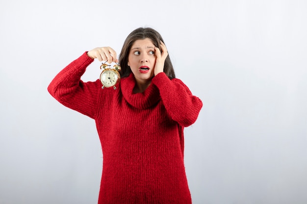 Jonge mooie donkerbruine vrouw die een wekker houdt die zich over witte achtergrond bevindt