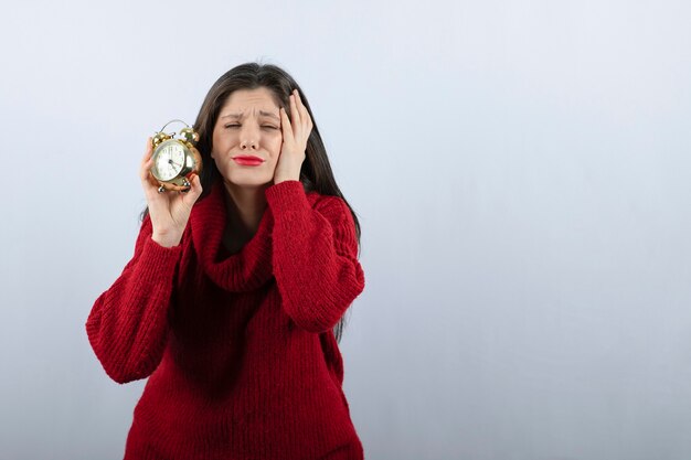 Jonge mooie donkerbruine vrouw die een wekker houdt die zich over witte achtergrond bevindt