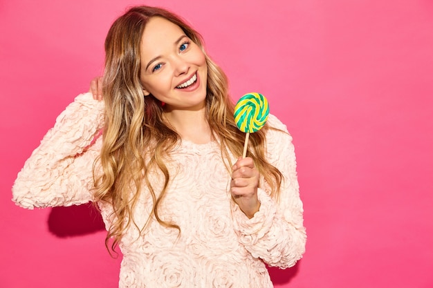 Jonge mooie glimlachende hipster vrouw in trendy zomerkleren. Het sexy onbezorgde vrouw stellen dichtbij roze muur. Positief model dat lolly eet