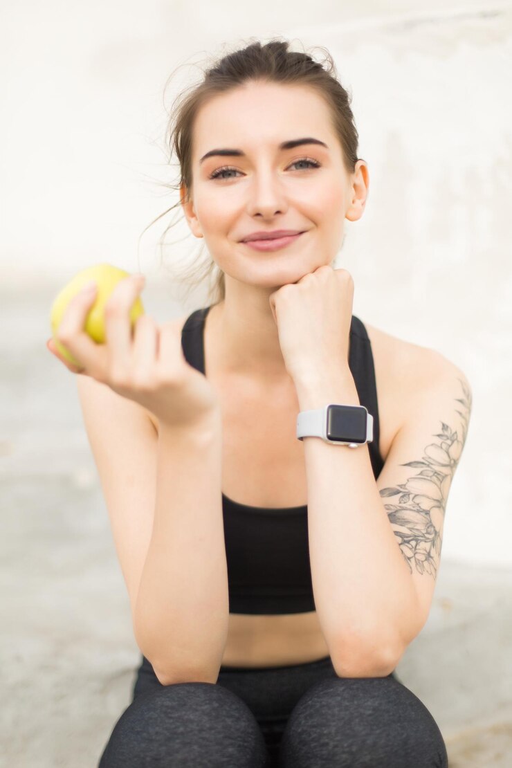 Gratis Foto jonge mooie glimlachende vrouw in sportieve top en legging die appel in de hand houdt en dromerig in de camera kijkt terwijl ze tijd buiten doorbrengt geïsoleerd