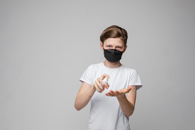 Jonge mooie Kaukasische tiener in wit t-shirt, zwarte spijkerbroek staat met zwarte medische masker desinfecteert zijn handen met anticepticum
