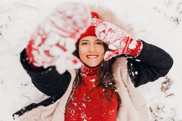 Jonge mooie lachende gelukkige vrouw in rode wanten en gebreide muts dragen winterjas liggend in park in de sneeuw, warme kleren, van bovenaf bekijken