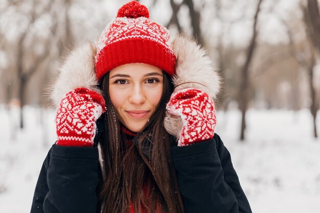 Jonge mooie lachende gelukkige vrouw in rode wanten en gebreide muts dragen winterjas met bont capuchon, wandelen in het park in de sneeuw, warme kleren