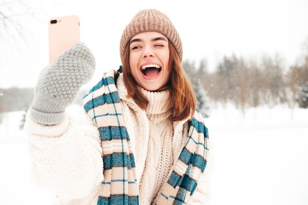 Jonge mooie lachende hipster vrouw in trendy warme kleding en sjaal. Zorgeloos vrouw poseren in de straat in het park. Positief puur model met plezier in de sneeuw. Genieten van winterse momenten. Selfie maken