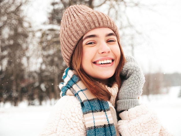 Jonge mooie lachende hipster vrouw in trendy warme kleding en sjaal. zorgeloos vrouw poseren in de straat in het park. positief puur model met plezier in de sneeuw. genieten van winterse momenten