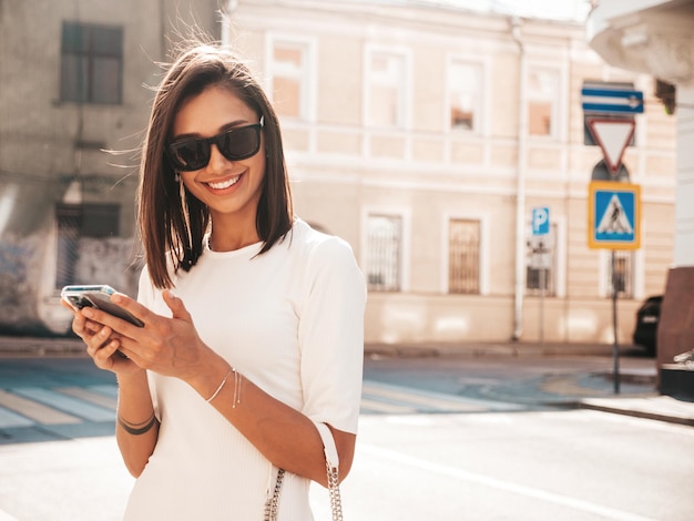 Jonge mooie lachende hipster vrouw in trendy zomerjurk Sexy zorgeloze vrouw die zich voordeed op de straat achtergrond bij zonsondergang Positief model smartphone-apps gebruiken kijken naar mobiel scherm