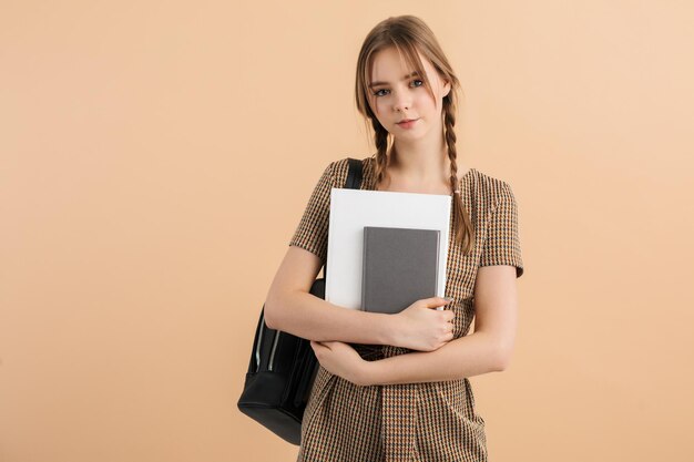 Jonge mooie lachende meisje met twee vlechten in tweed jumpsuit met zwarte rugzak op schouder notitieblokken in handen houden terwijl gelukkig in de camera kijken over beige achtergrond