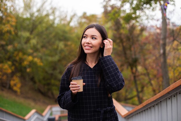 Jonge mooie lachende vrouw in jas met koffie om vrolijk opzij te kijken in het park