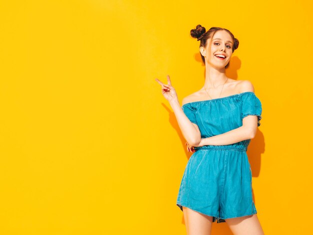 Jonge mooie lachende vrouw in trendy zomer jeans overall Sexy zorgeloze vrouw met twee hoorns kapsel poseren in de buurt van gele muur in studio Positief model plezier geïsoleerd