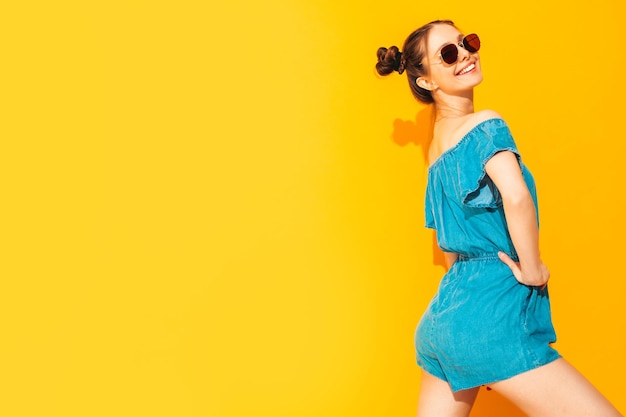 Jonge mooie lachende vrouw in trendy zomer jeans overall Sexy zorgeloze vrouw met twee hoorns kapsel poseren in de buurt van gele muur in studio Positief model plezier In zonnebril geïsoleerd