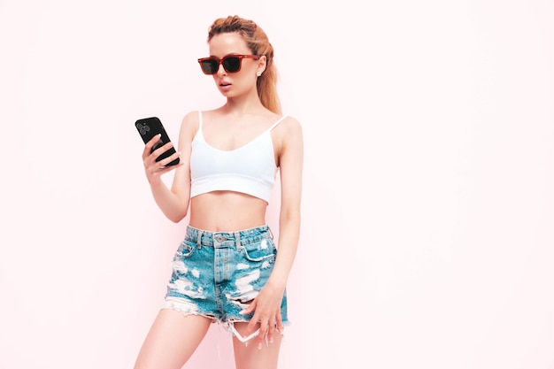 Jonge mooie lachende vrouw in trendy zomer jeans shorts Sexy zorgeloze blonde vrouw poseren in de buurt van muur in studio Positief model plezier Holding smartphone en het gebruik van mobiele apps