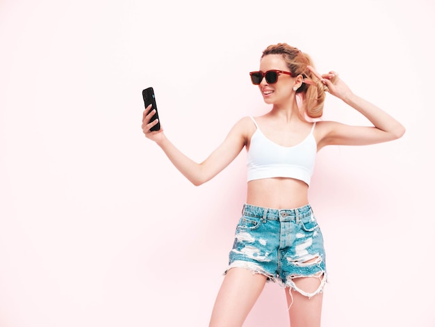 Jonge mooie lachende vrouw in trendy zomer jeans shorts sexy zorgeloze blonde vrouw poseren in de buurt van muur in studio positief model plezier vrolijk en gelukkig in zonnebril geïsoleerd nemen selfie