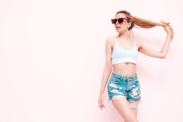 Jonge mooie lachende vrouw in trendy zomer jeans shorts Sexy zorgeloze blonde vrouw poseren in de buurt van muur in studio Positief model plezier vrolijk en gelukkig in zonnebril geïsoleerd