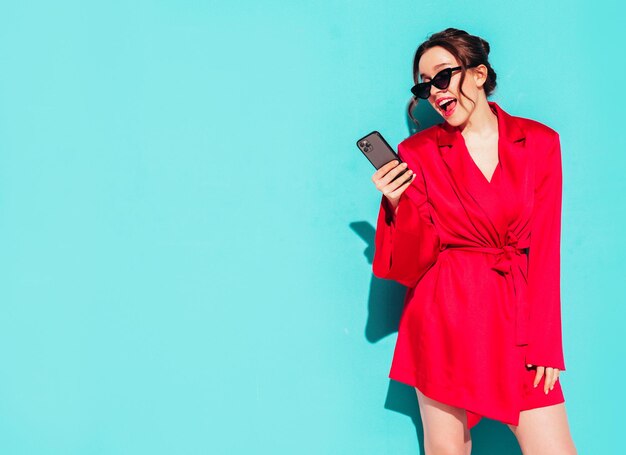 Jonge mooie lachende vrouw in trendy zomer rode jurk Sexy zorgeloze vrouw poseren in de buurt van blauwe muur in studio Positief model plezier kijken naar smartphone scherm en het gebruik van apps In zonnebril