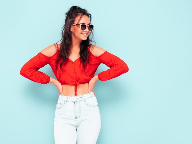 Jonge mooie lachende vrouw in trendy zomer rode top en jeans kleding Sexy zorgeloze vrouw poseren in de buurt van blauwe muur in studio Positieve brunette model plezier vrolijk en gelukkig In zonnebril