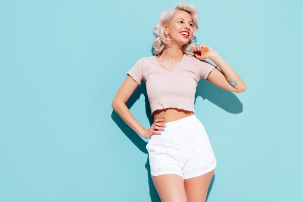 Jonge mooie lachende vrouw in trendy zomer shorts en topkleren Sexy zorgeloze vrouw poseren in de buurt van blauwe muur in studio Positieve blond model plezier en gek vrolijk en gelukkig