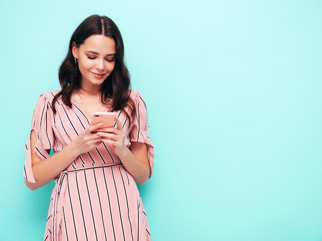 Jonge mooie lachende vrouw in trendy zomerjurk Sexy zorgeloze vrouw poseren in de buurt van blauwe muur in studio Positieve brunette model kijken naar mobiele telefoon scherm Holding smartphone en het gebruik van apps