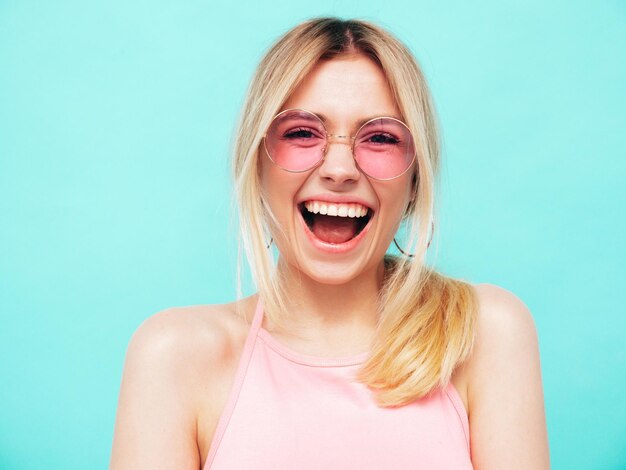 Jonge mooie lachende vrouw in trendy zomerkleren Sexy zorgeloze vrouw poseren in de buurt van blauwe muur in studio Positief model plezier vrolijk en gelukkig In zonnebril schreeuwen en schreeuwen