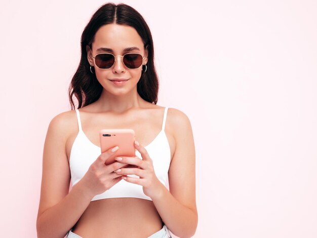 Jonge mooie lachende vrouw in trendy zomerkleren Sexy zorgeloze vrouw poseren in de buurt van roze muur in studio Positieve brunette model kijken naar mobiele telefoon scherm Holding smartphone en het gebruik van apps