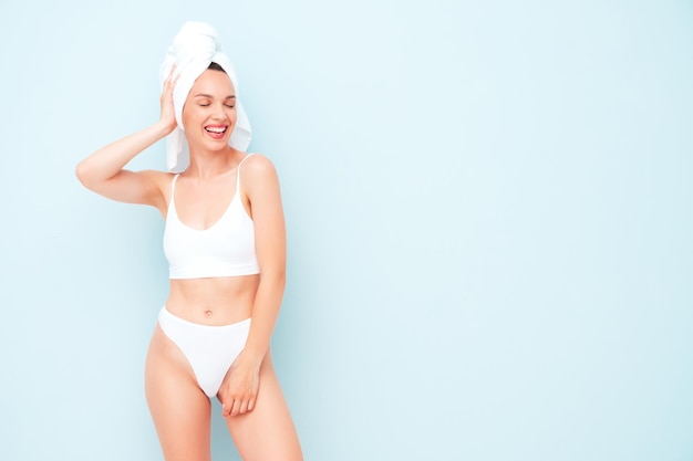 Jonge mooie lachende vrouw in witte lingerie. Sexy zorgeloos model in ondergoed en handdoek op hoofd poseren in de buurt van lichtblauwe muur in studio. Positieve en gelukkige vrouw die van de ochtend geniet