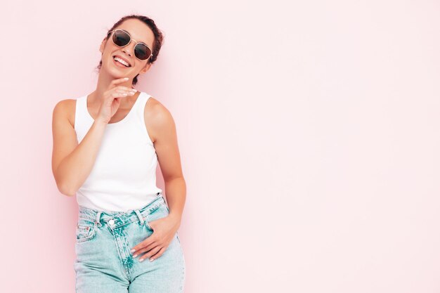 Jonge mooie lachende vrouw in witte zomer tshirt en jeans Sexy zorgeloze vrouw poseren in de buurt van roze muur in studio Positieve model plezier en gek vrolijk en gelukkig In zonnebril