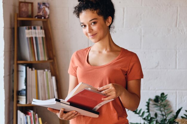 Jonge mooie lachende vrouw met donker krullend haar in t-shirt met boeken in handen die gelukkig in de camera kijken en in de buurt van een boekenplank staan in een gezellig huis