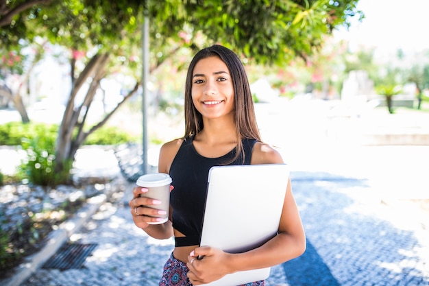 Jonge mooie Latijns-student met laptop en kopje koffie studeren in het park. Meisje loopt in het park met een grote glimlach en houdt laptop.