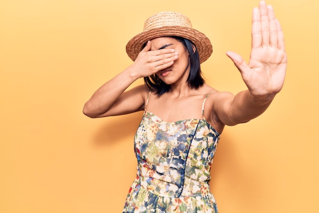 Jonge mooie latijns-vrouw die een zomerhoed draagt die de ogen bedekt met handen en een stopgebaar doet met droevige en angstuitdrukking. beschaamd en negatief concept.