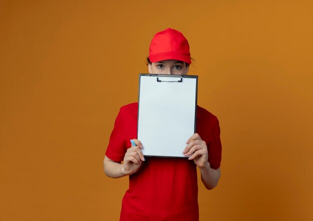 Jonge mooie levering meisje in rood uniform en cap Klembord pen houden en kijken naar camera van achter Klembord geïsoleerd op een oranje achtergrond met kopie ruimte