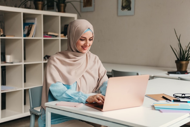 Jonge mooie moderne moslimvrouw in hijab die op laptop in kantoorruimte werkt, online onderwijs education