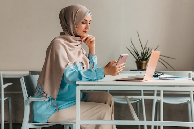 Jonge mooie moderne moslimvrouw in hijab die op laptop in kantoorruimte werkt, online onderwijs education