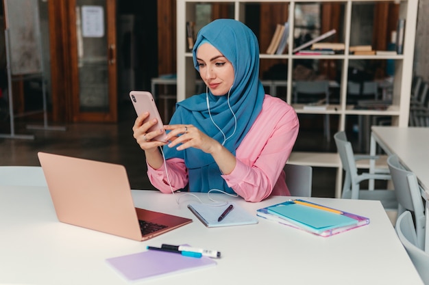 Jonge mooie moderne moslimvrouw in hijab die op laptop in kantoorruimte werkt, online onderwijs education