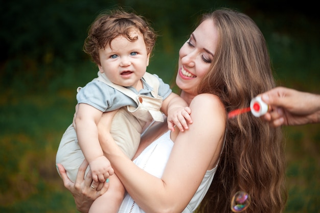 Jonge mooie moeder die haar kleine peuterzoon koestert tegen groen gras. Gelukkige vrouw met haar babyjongen op een de zomer zonnige dag. Familie die op de weide loopt.