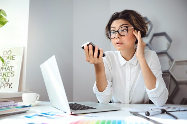 Jonge mooie onderneemster die op telefoon op werkplaats in bureau kijkt.