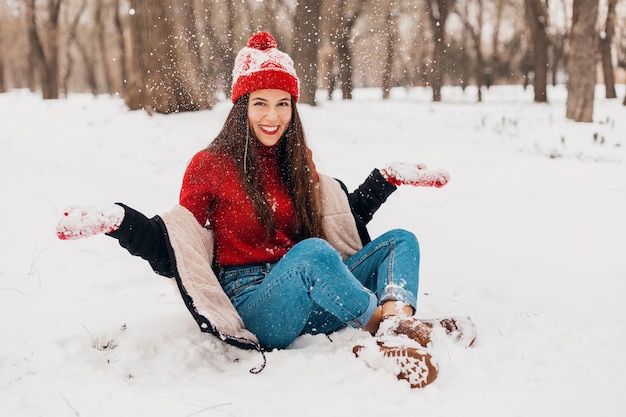 Gratis foto jonge mooie openhartige lachende gelukkige vrouw in rode wanten en gebreide muts dragen zwarte jas wandelen spelen in park in de sneeuw, warme kleren, plezier