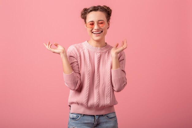 Jonge mooie oprechte lachende vrouw in roze trui en zonnebril geïsoleerd op roze studio achtergrond, openhartige glimlach