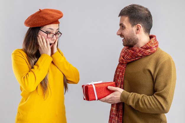 Jonge mooie paar gelukkige man die een cadeau geeft voor zijn lachende en verbaasde vriendin in baret gelukkig verliefd samen vieren valentijnsdag staande op witte achtergrond