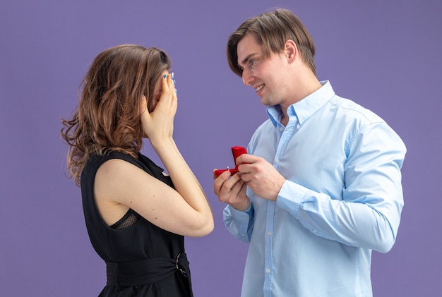 Jonge mooie paar gelukkige man die een voorstel doet met verlovingsring in rode doos voor zijn mooie vriendin die ogen bedekt met handen tijdens valentijnsdag staande over blauwe achtergrond