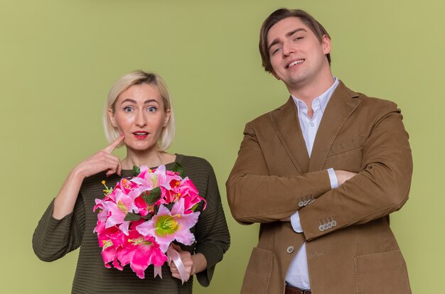 Jonge mooie paar gelukkige man en vrouw met boeket bloemen kijken voorkant glimlachend vrolijk vieren internationale Vrouwendag staande over groene muur