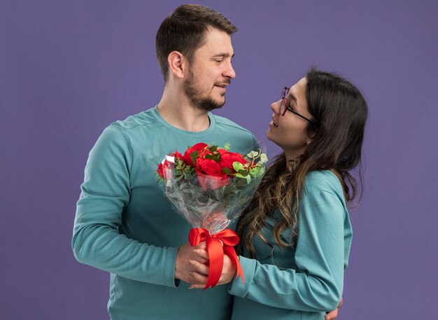 Jonge mooie paar in blauwe casual kleding man en vrouw met een boeket rozen kijken elkaar gelukkig verliefd samen permanent over paarse muur