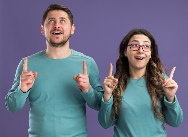 Jonge mooie paar in blauwe vrijetijdskleding man en vrouw glimlachend omhoog kijken vrolijk wijzend met wijsvingers omhoog staan over paarse muur