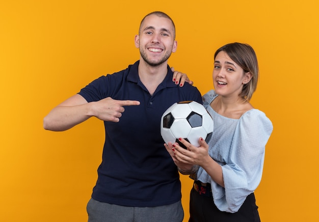 Jonge mooie paar in casual kleding lachende vrouw met voetbal terwijl haar lachende vriendje met wijsvinger wijzend op bal staande over oranje muur