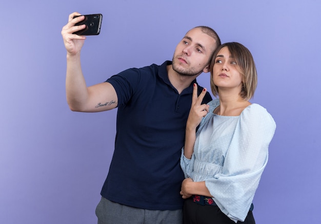 Jonge mooie paar man en vrouw gelukkig en positief doen selfie samen met smartphone met v-teken staande over blauwe muur